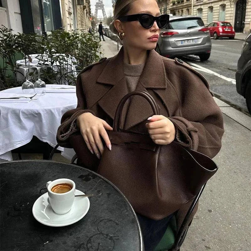 Elegant brown woolen overcoat, double-breasted, worn by woman sitting at outdoor cafe table with coffee.